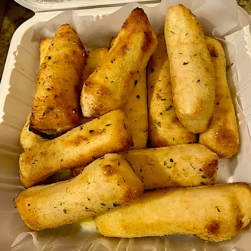 Golden-brown garlic breadsticks, seasoned with herbs, fill a white takeout container from Pizza Pizazz.