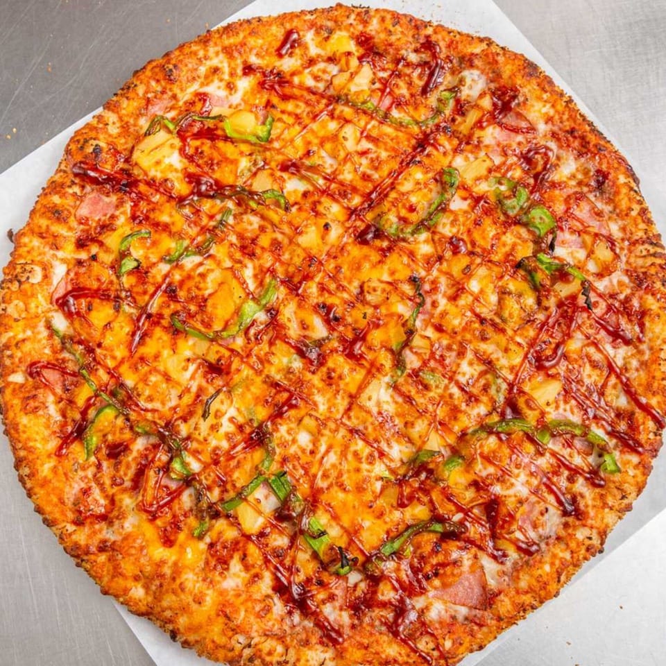 A whole Pizza Pizazz pizza, topped with cheese, pineapple, green peppers, and a BBQ sauce drizzle.