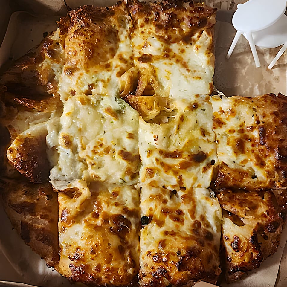 A close-up of a square-cut, cheesy white pizza with a golden-brown crust in a pizza box.